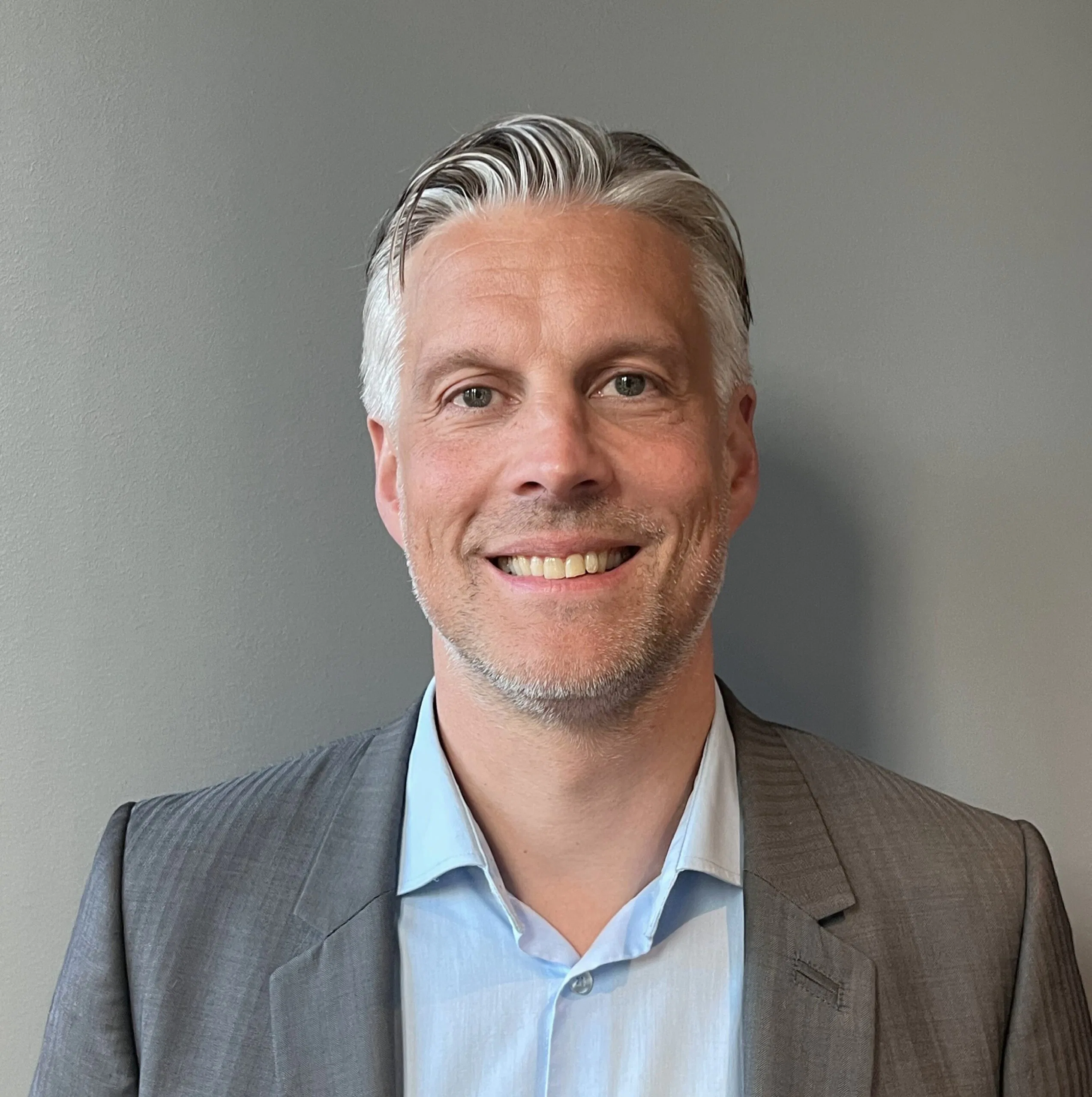 Smiling adult man wearing a gray blazer and light blue shirt, photographed against a plain gray wall from the chest up.