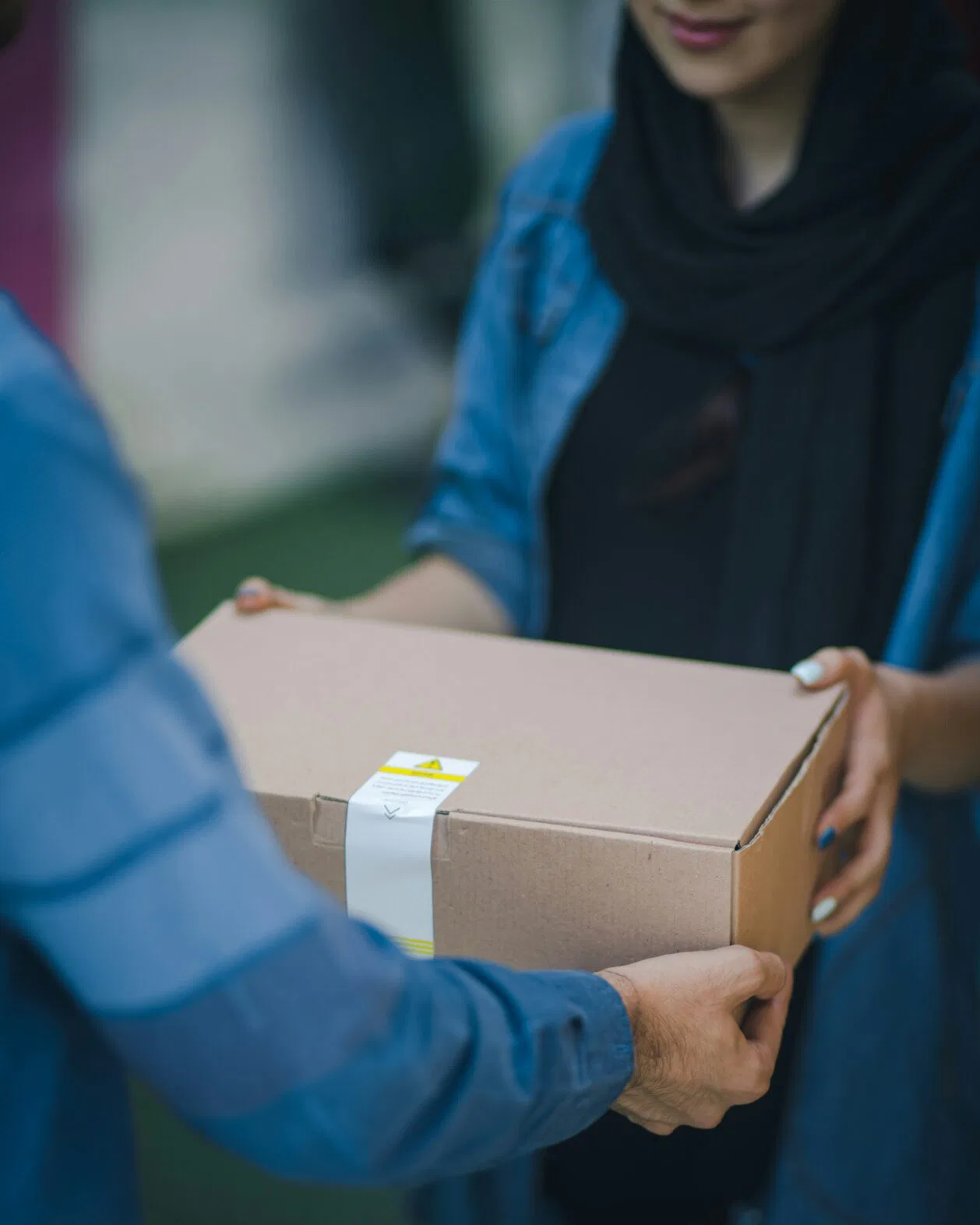 Two people pass a cardboard box taped shut outdoors. (handoff)